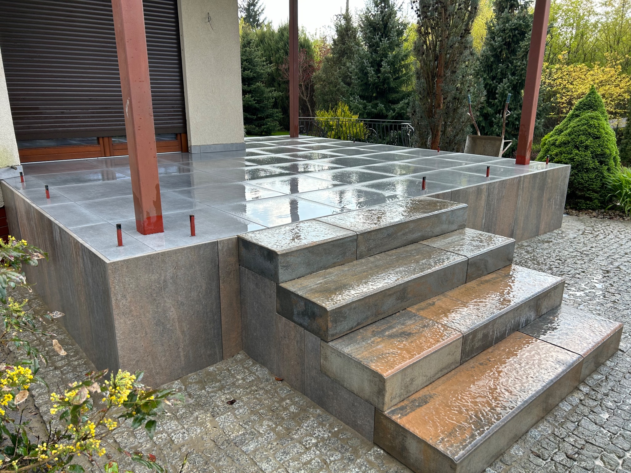 Nowoczesny taras z szarych, kwadratowych płyt, z niskimi schodami wykonanymi z kostki o zróżnicowanej kolorystyce, otoczony zielenią i kostką brukową na podwórku, lekko mokry po deszczu.