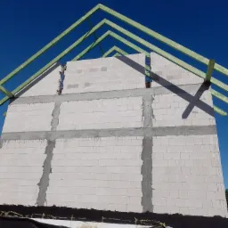 Wznoszenie ściany z białych bloczków z widocznymi wzmocnieniami pionowymi i poziomymi oraz konstrukcją dachu z zielonych belek na tle intensywnego, błękitnego nieba.