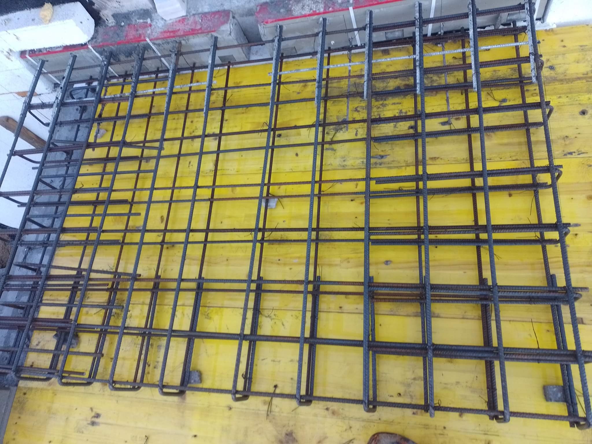 Zbrojenie fundamentu ułożone na żółtej płycie szalunkowej, widoczne pionowe pręty zagięte u podstawy.