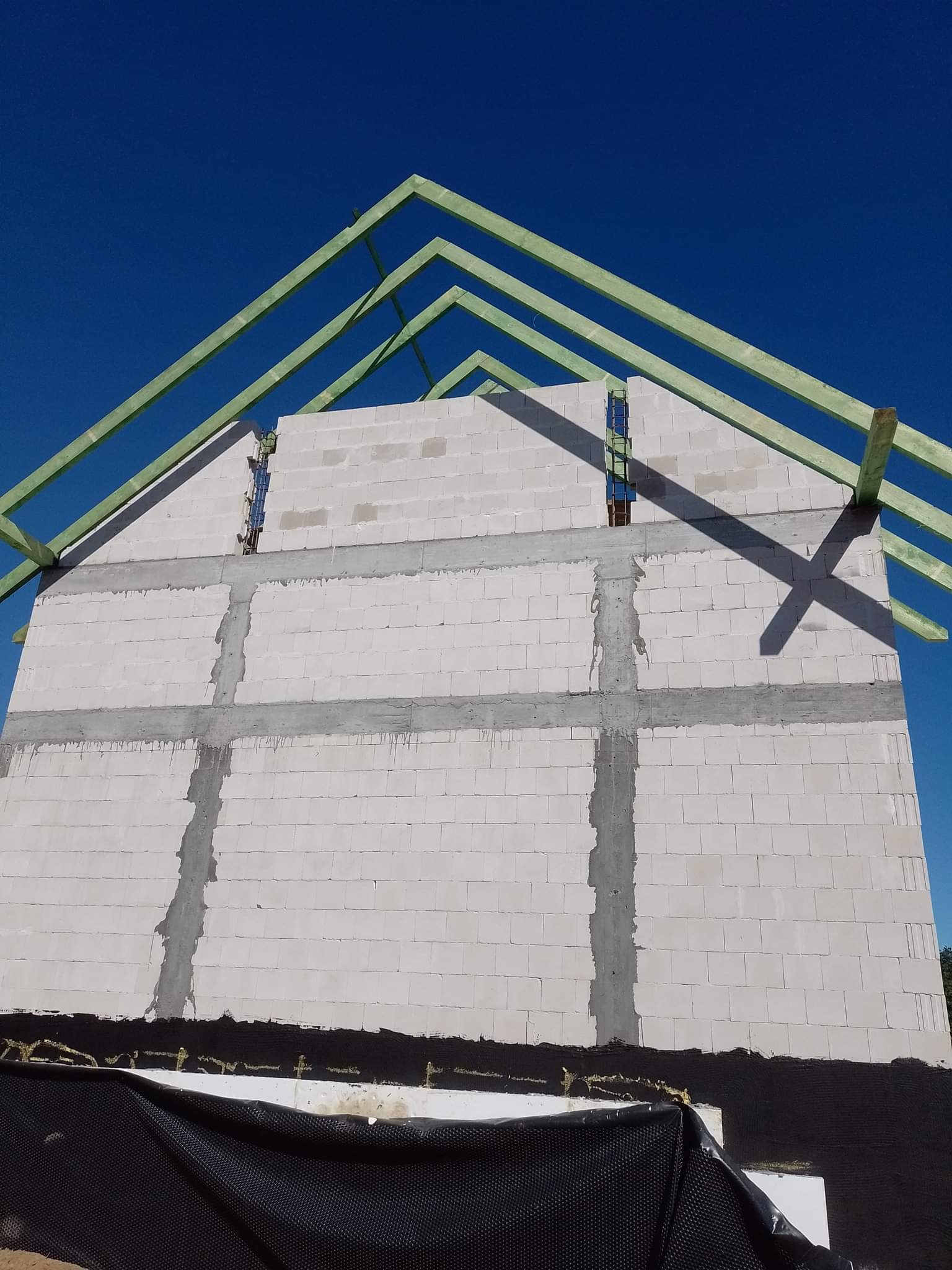 Wznoszenie ściany z białych bloczków z widocznymi wzmocnieniami pionowymi i poziomymi oraz konstrukcją dachu z zielonych belek na tle intensywnego, błękitnego nieba.