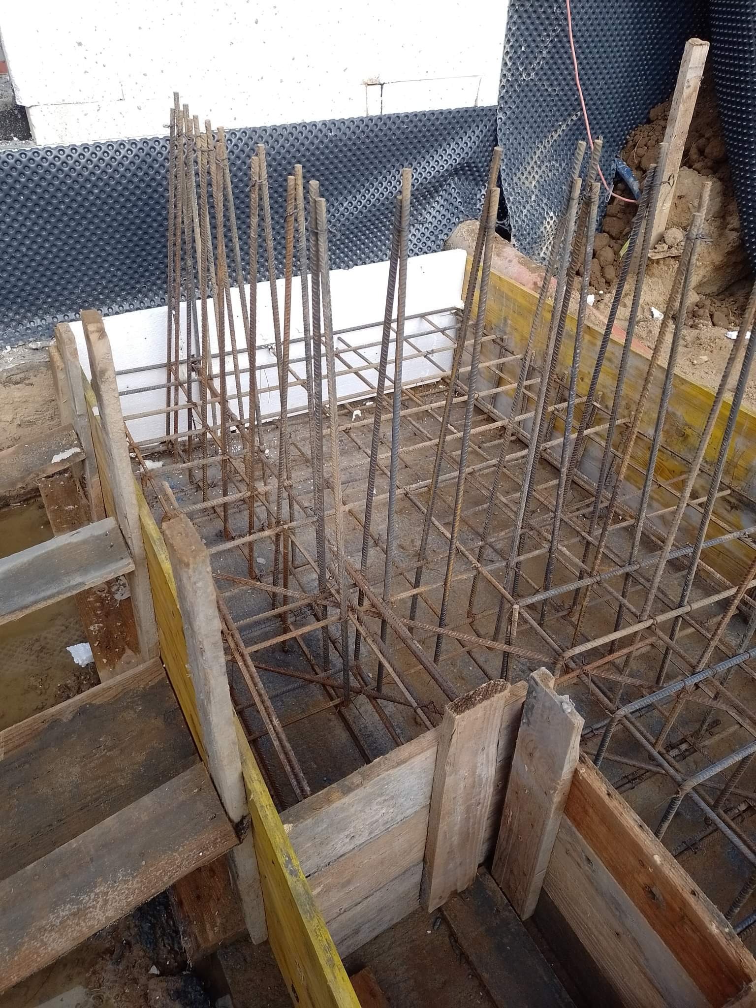 Zbrojenie fundamentu w drewnianym szalunku, z widoczną izolacją pionową i fragmentem ściany z białych bloczków.