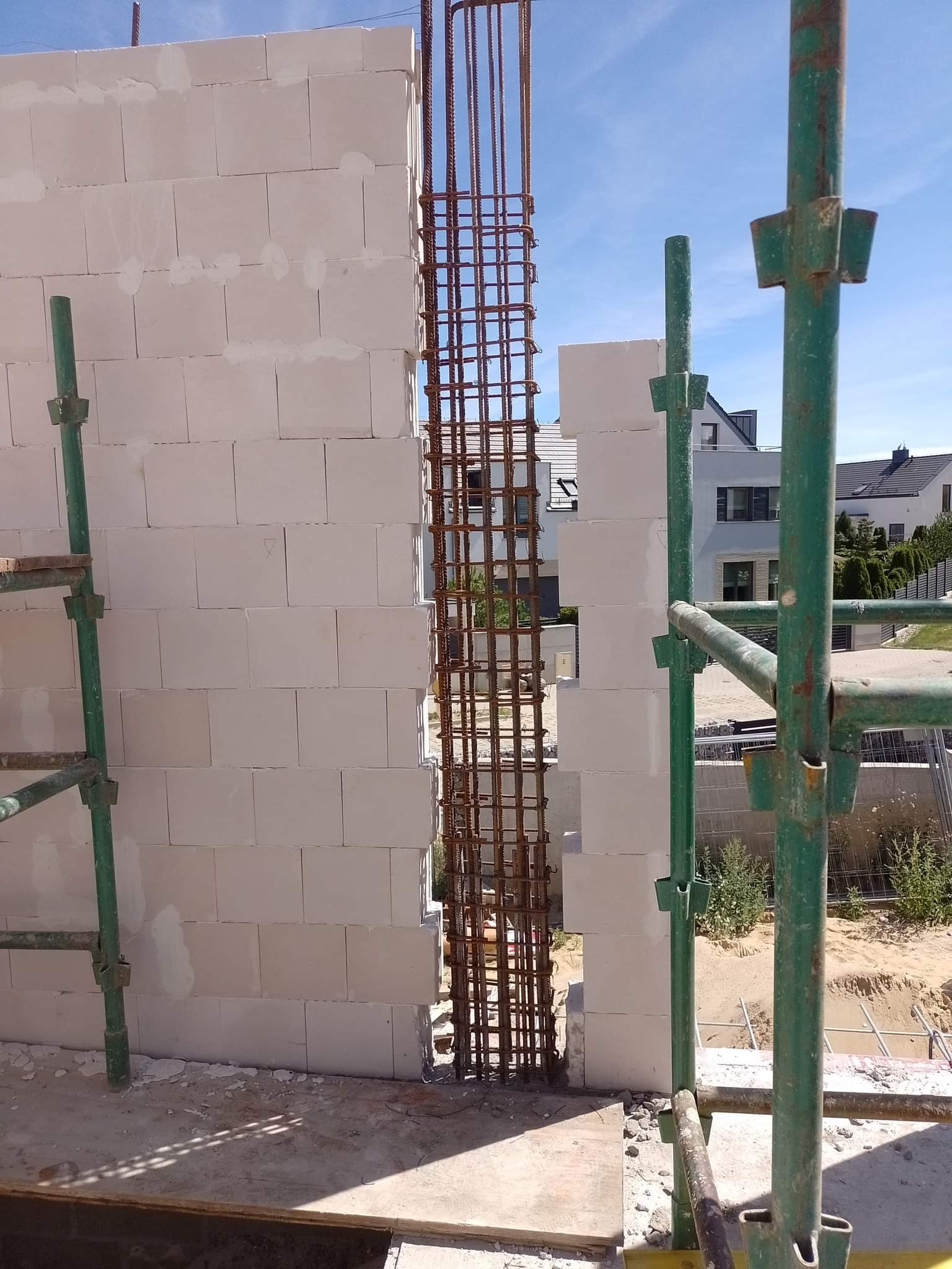 Wznoszenie ściany z betonu komórkowego Ytong z przerwą na zbrojenie pionowe, widoczne zielone rusztowanie i fragment zabudowy jednorodzinnej w tle.