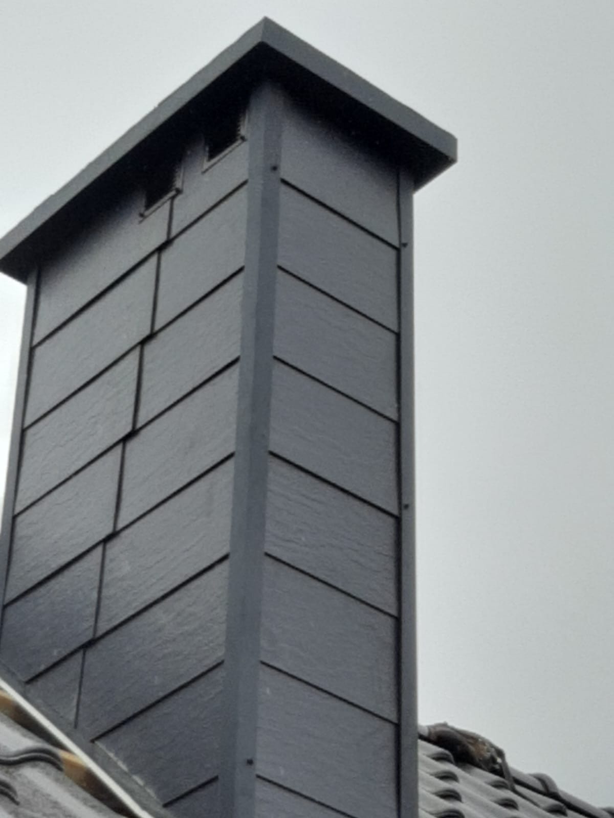 Szary komin obłożony łupkiem, widoczny z dołu, na tle pochmurnego nieba.