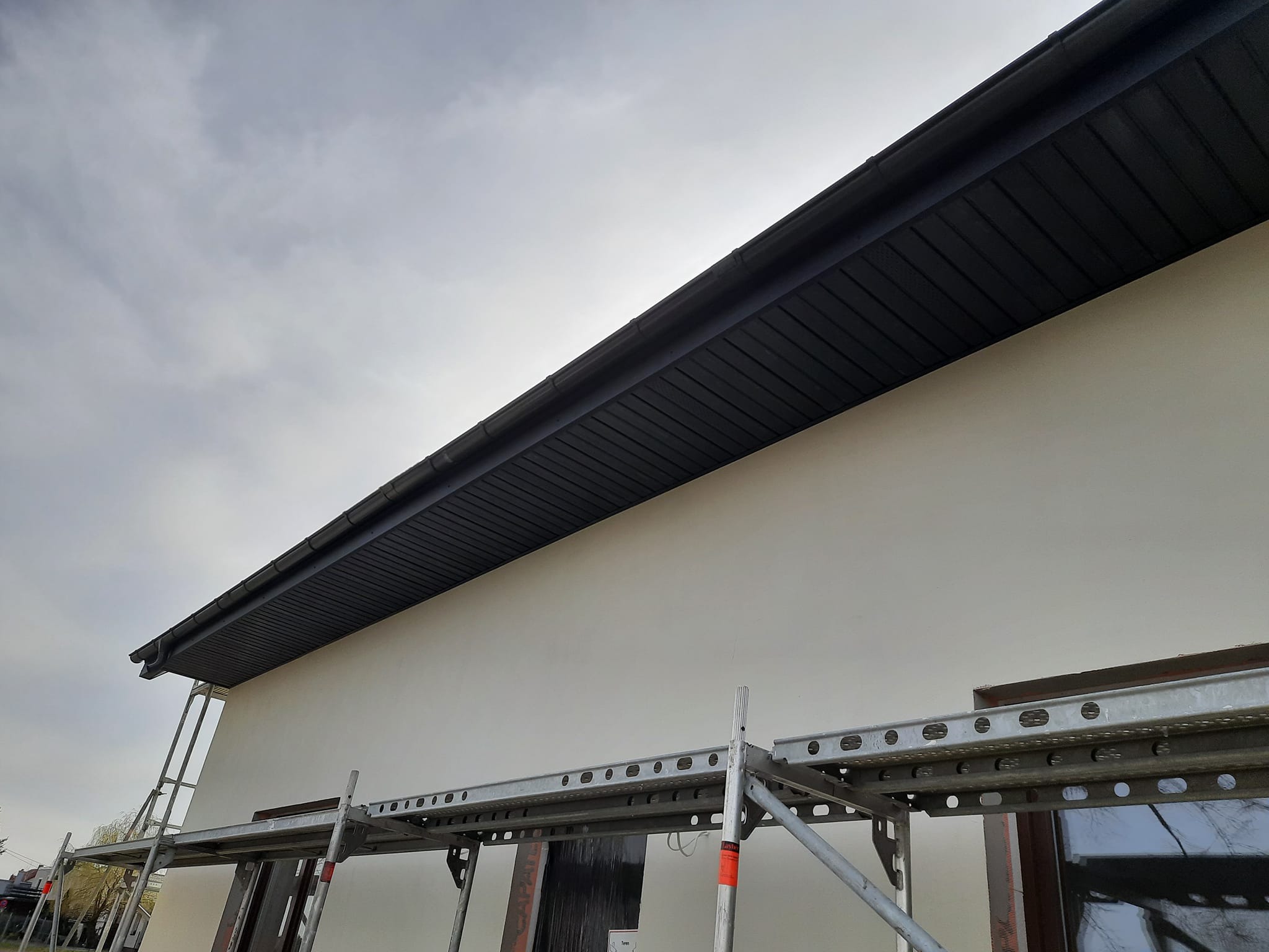 Szare podbitki dachowe zamontowane na budynku, widoczne rusztowanie z boku, pochmurne niebo w tle.