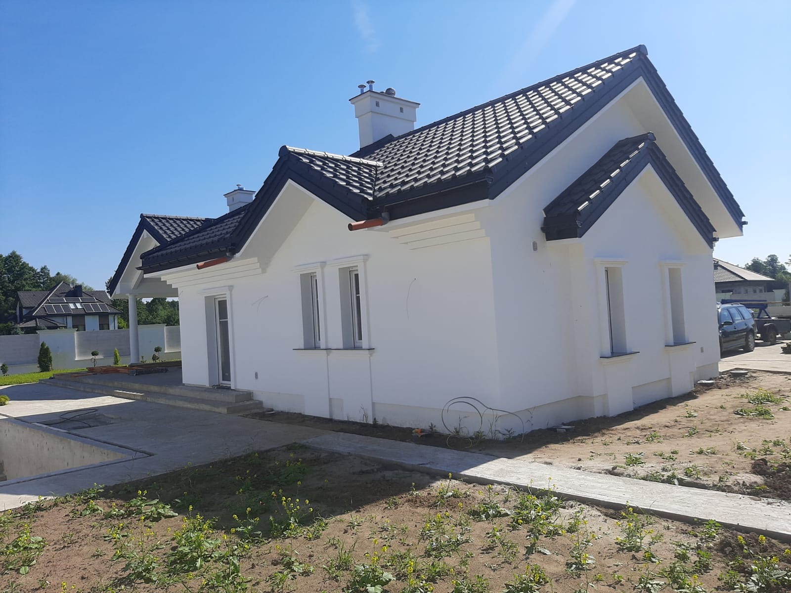 Nowoczesny dom jednorodzinny z czarną dachówką karpiówką i białymi ścianami, widok z boku na ogród w trakcie aranżacji.