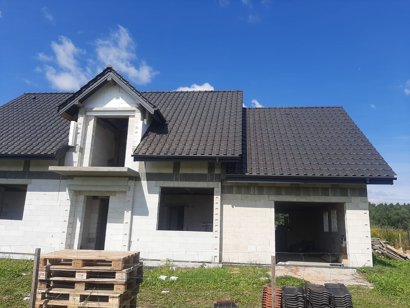 Dom w trakcie budowy z nowym, ciemnoszarym dachem z dachówki ceramicznej i zamontowanymi rynnami, białe ściany z betonu komórkowego, palety i dachówki na trawie.