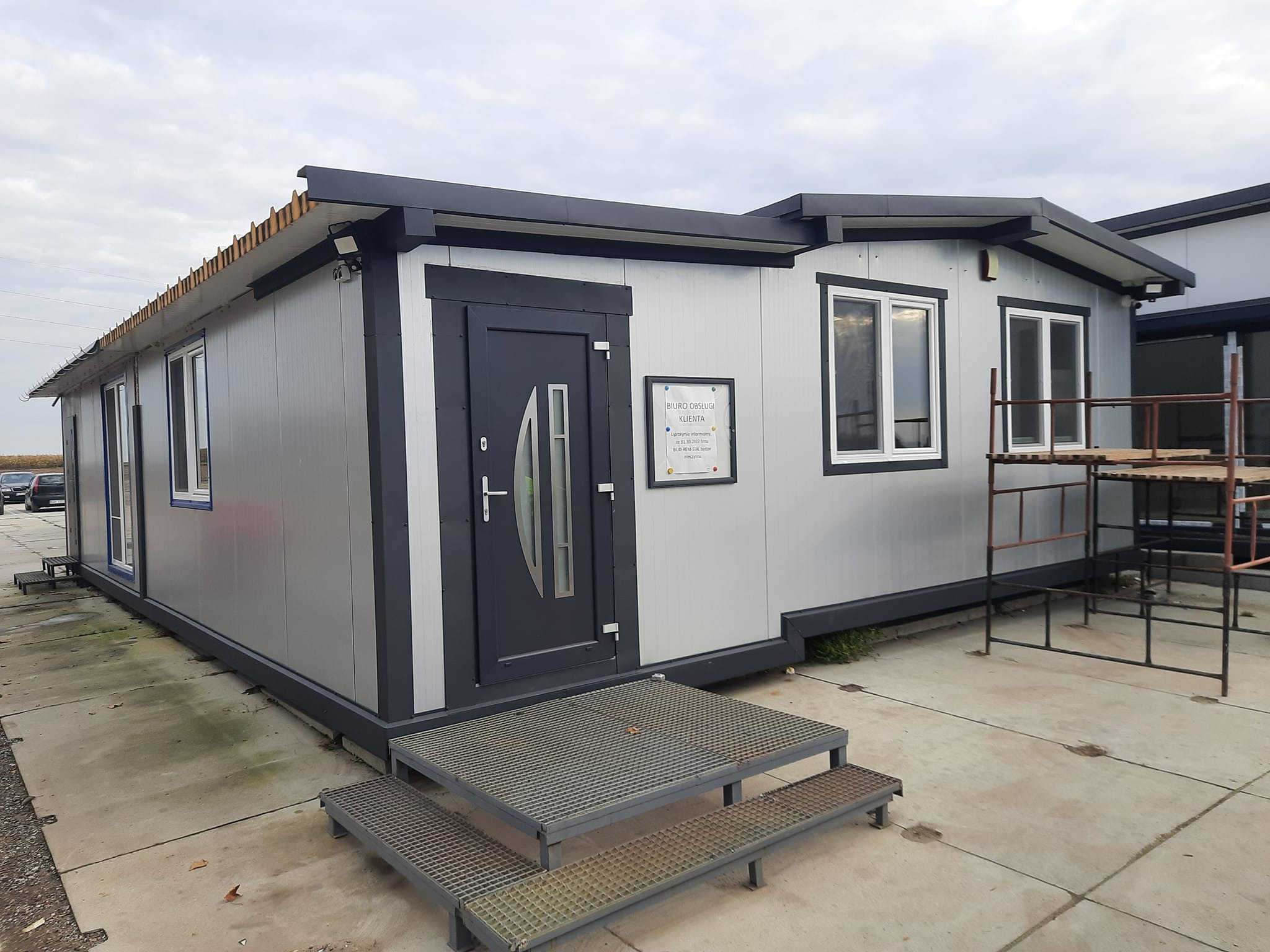 Nowoczesny modułowy budynek biurowy z płaskim dachem, szarą elewacją i metalowymi schodami wejściowymi, tabliczka informacyjna 'Biuro Obsługi Klienta' przy drzwiach.