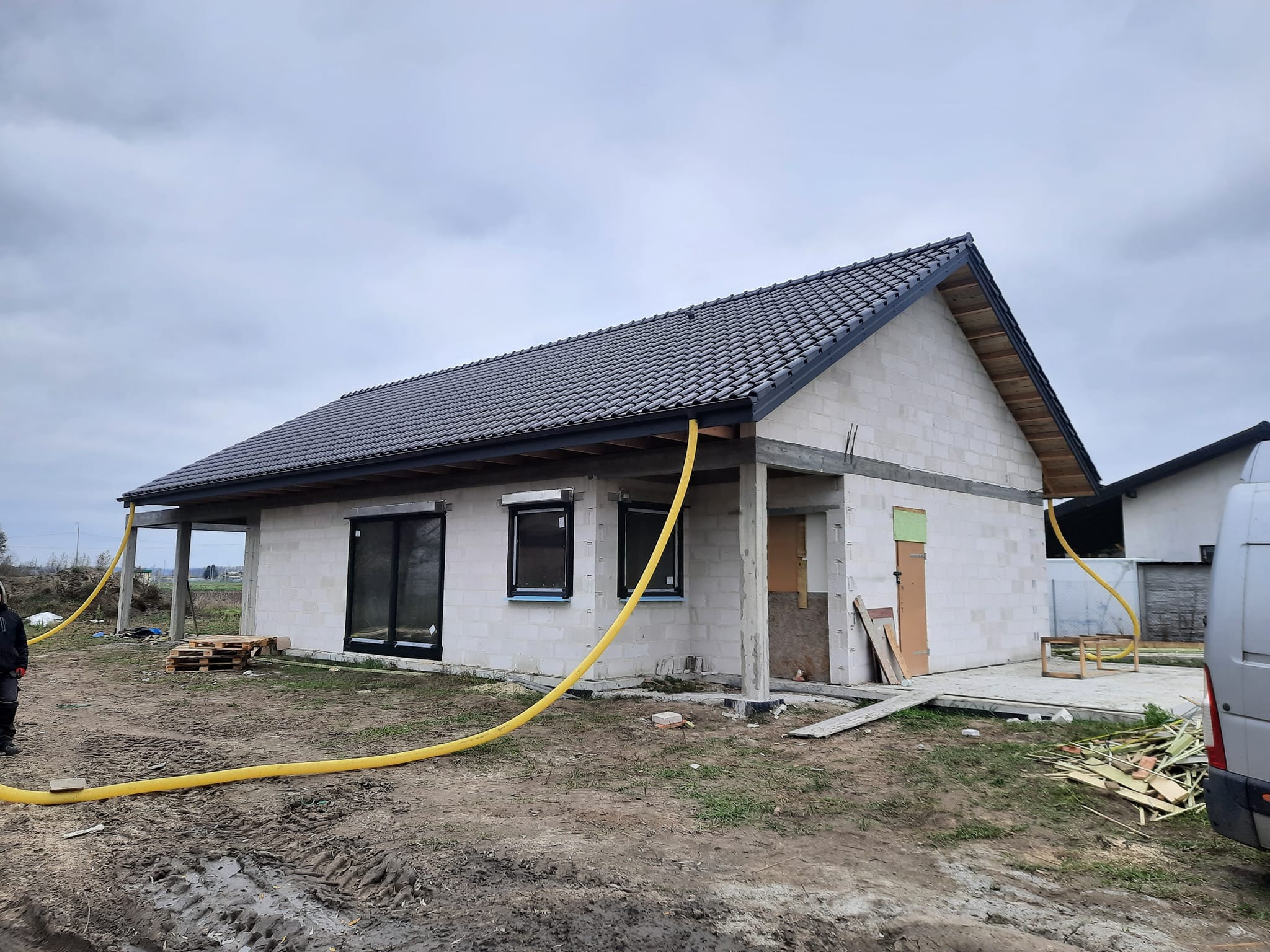 Surowy stan domu jednorodzinnego z szarej cegły, z czarną dachówką, widoczne żółte węże doprowadzające media, teren wokół domu w trakcie prac budowlanych, pochmurne niebo.