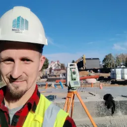 Mężczyzna w kasku z logo firmy i kamizelce odblaskowej robi selfie na tle budowy z niwelatorem na statywie i koparką w oddali.