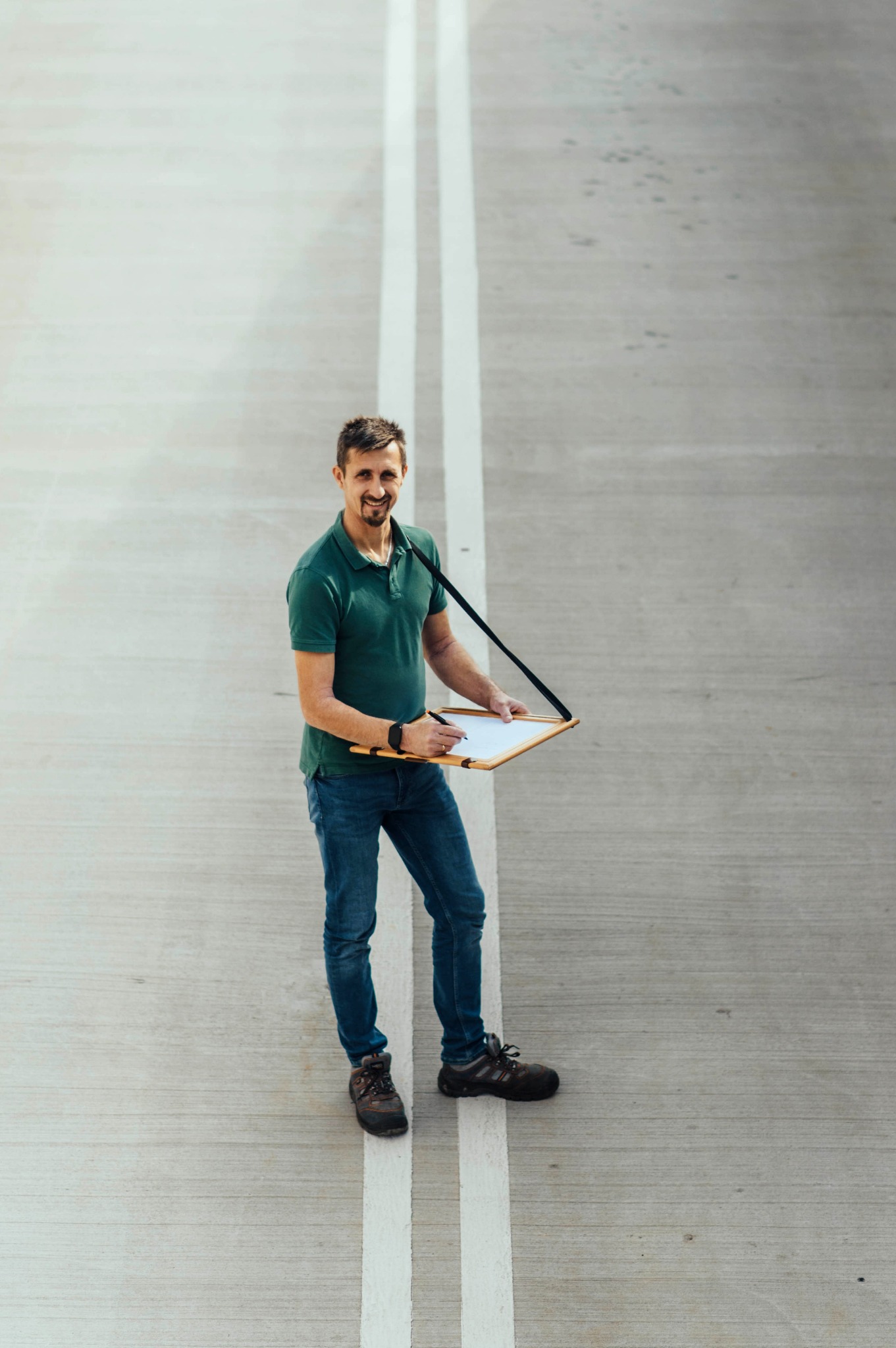 Uśmiechnięty mężczyzna w zielonej koszulce polo i jeansach, stojący na betonowej powierzchni z białymi liniami, trzymający deskę z papierem i długopisem, widziany z góry.