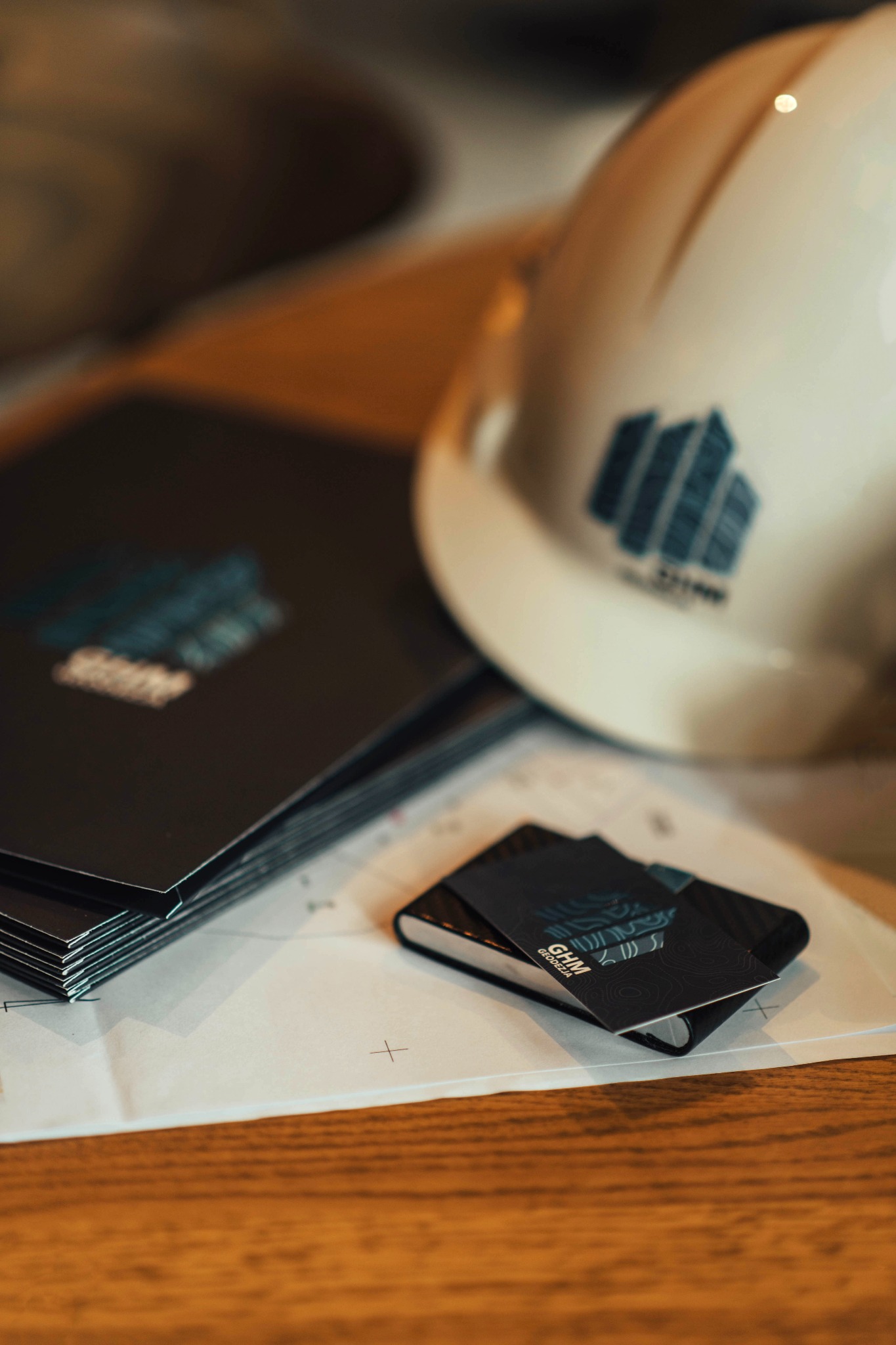 Biały kask budowlany z logo firmy na planie architektonicznym, obok leżą wizytówki i teczka z logo firmy.