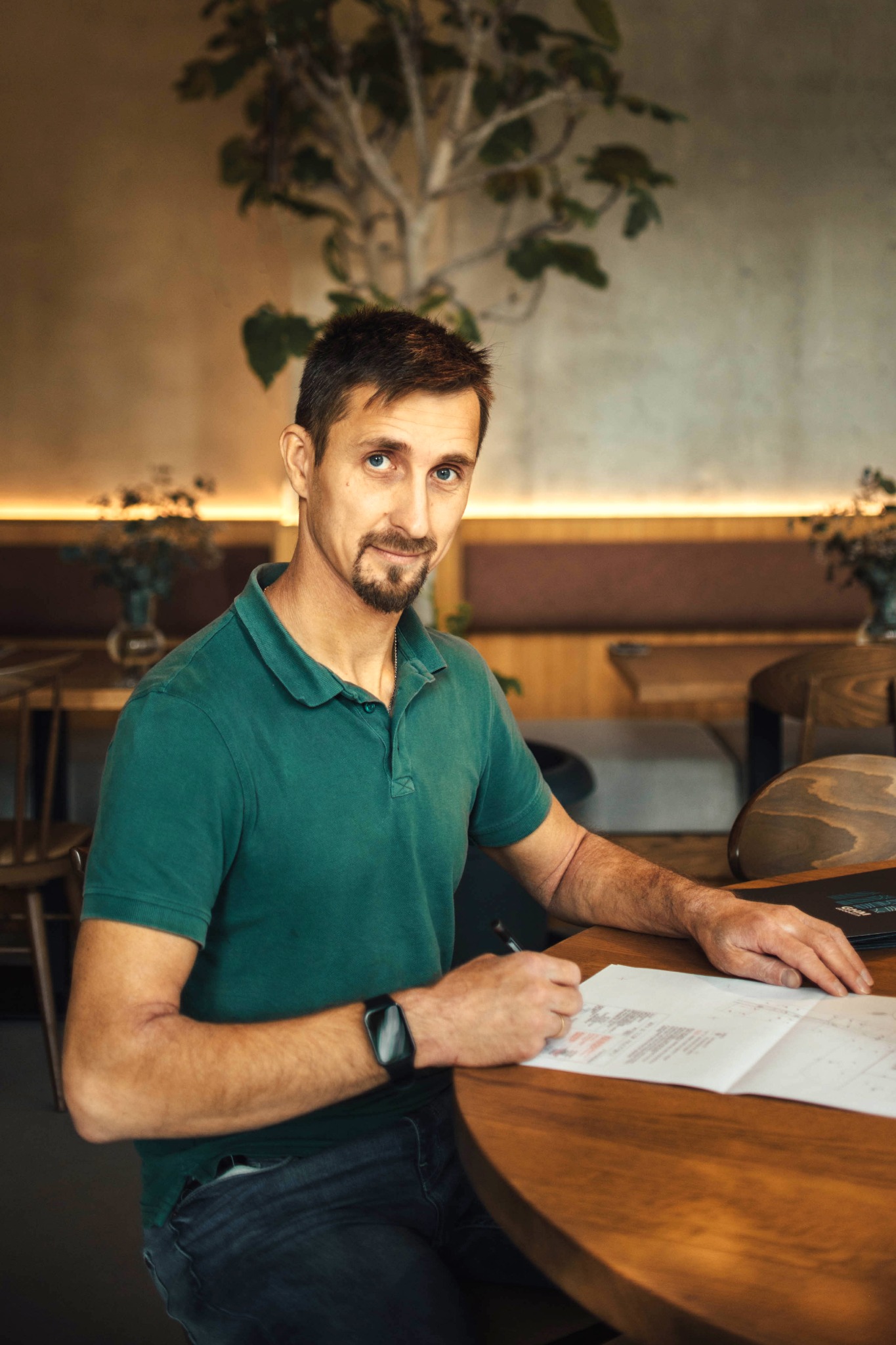 Mężczyzna z lekkim uśmiechem, w zielonej koszulce polo, siedzi przy okrągłym, drewnianym stole, analizując rozłożony plan budowlany i robi notatki czarnym długopisem. W tle widać rośliny doniczkowe...