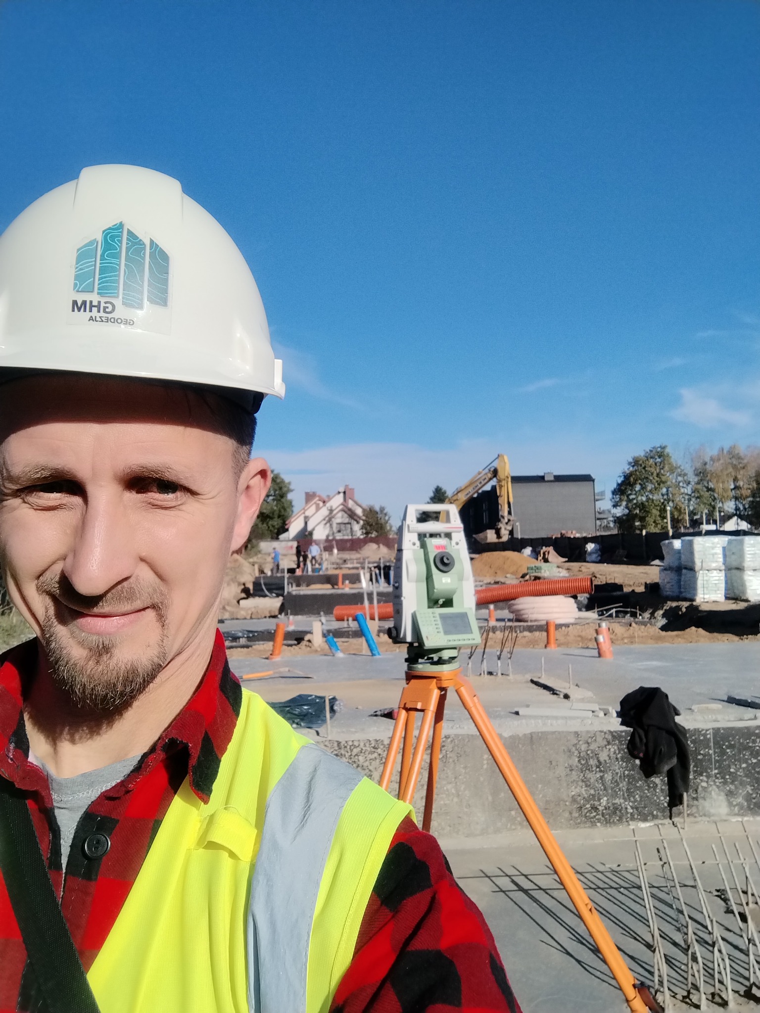 Mężczyzna w kasku z logo firmy i kamizelce odblaskowej robi selfie na tle budowy z niwelatorem na statywie i koparką w oddali.