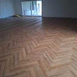 Jasne, dębowe panele ułożone we wzór jodełki francuskiej w przestronnym, jasnym salonie z dużym oknem tarasowym.