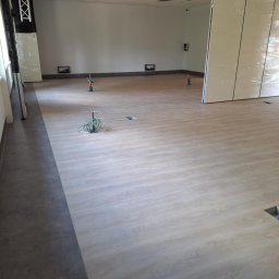 JARMAT Jarosław Wojnar - Jasne panele podłogowe w przestronnym, jasnym wnętrzu biurowym. Widoczne kable i otwory w podłodze, sugerujące prace wykończeniowe.