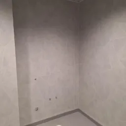 Wykończone wnętrze z szarą, geometryczną wykładziną ścienną i podłogową. Widoczne metalowe elementy sufitu podwieszanego i odpływ podłogowy.