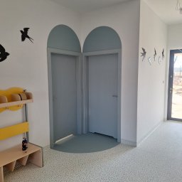 JARMAT Jarosław Wojnar - Jasne wnętrze z dwoma szarymi drzwiami z łukowatymi nadprożami, dekoracjami ptaków na ścianach i podłogą w kropki. Widoczny fragment wieszaka z ławką.