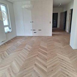 JARMAT Jarosław Wojnar - Jasny parkiet ułożony we wzór jodełki francuskiej w przestronnym, nowoczesnym wnętrzu. Widoczne gniazdka elektryczne i okno z lewej strony.