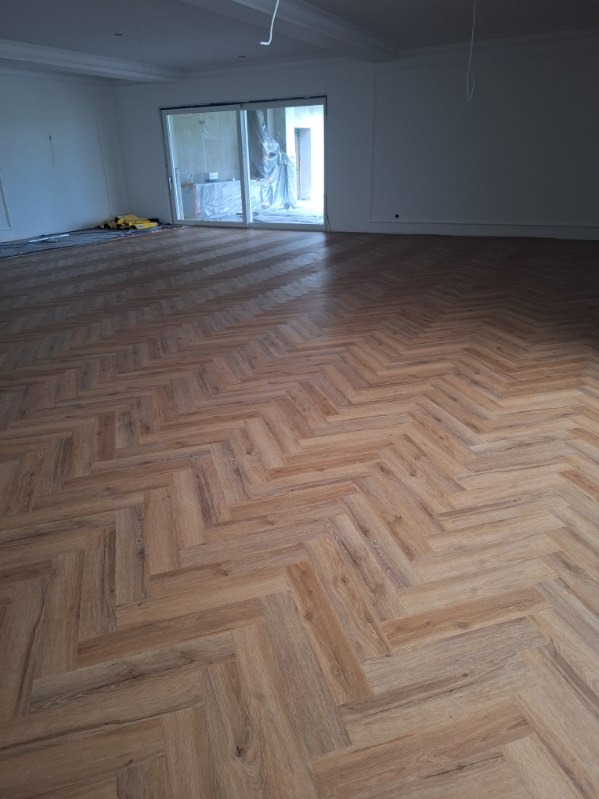 Jasne, dębowe panele ułożone we wzór jodełki francuskiej w przestronnym, jasnym salonie z dużym oknem tarasowym.