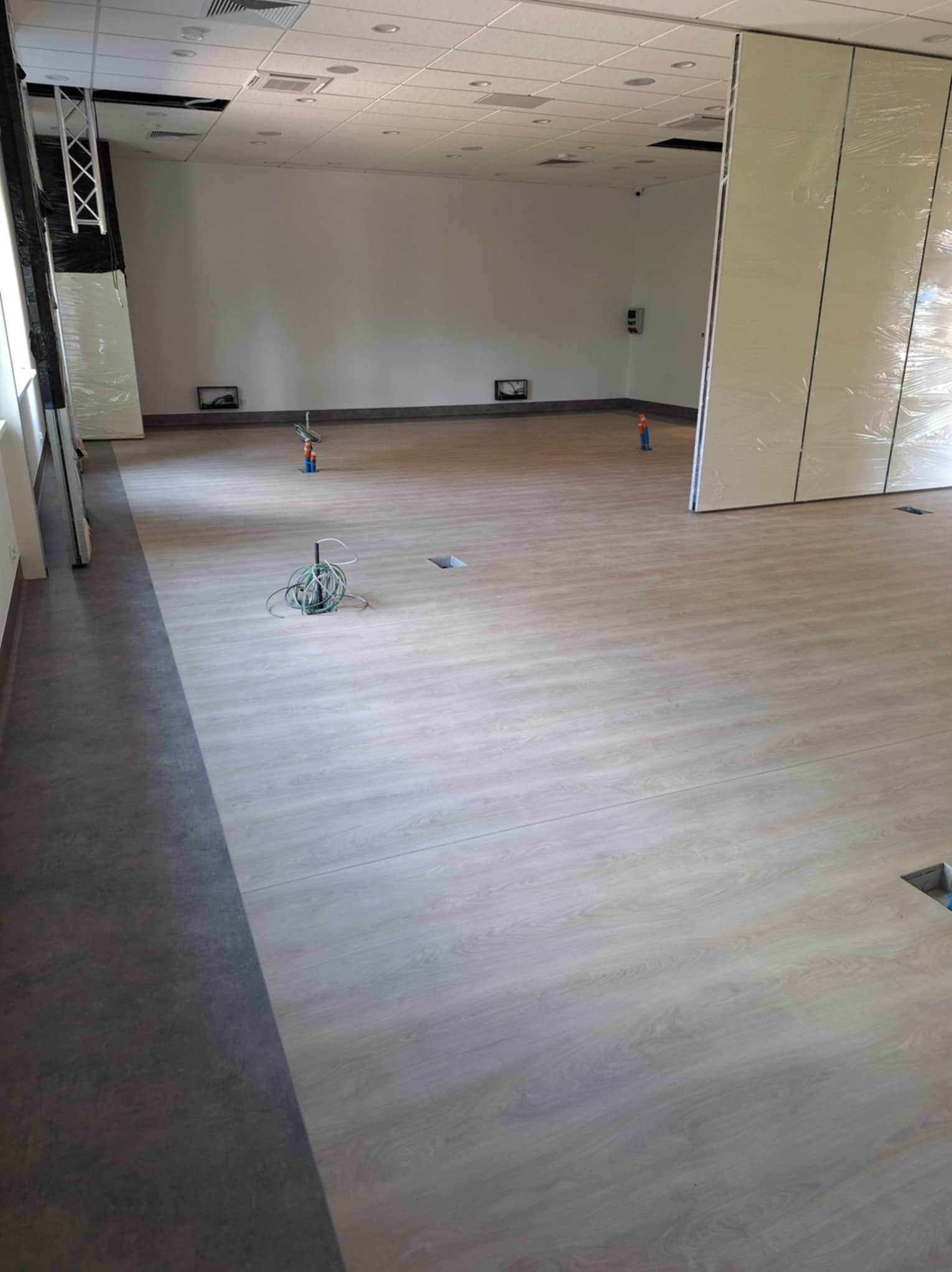 Jasne panele podłogowe w przestronnym, jasnym wnętrzu biurowym. Widoczne kable i otwory w podłodze, sugerujące prace wykończeniowe.