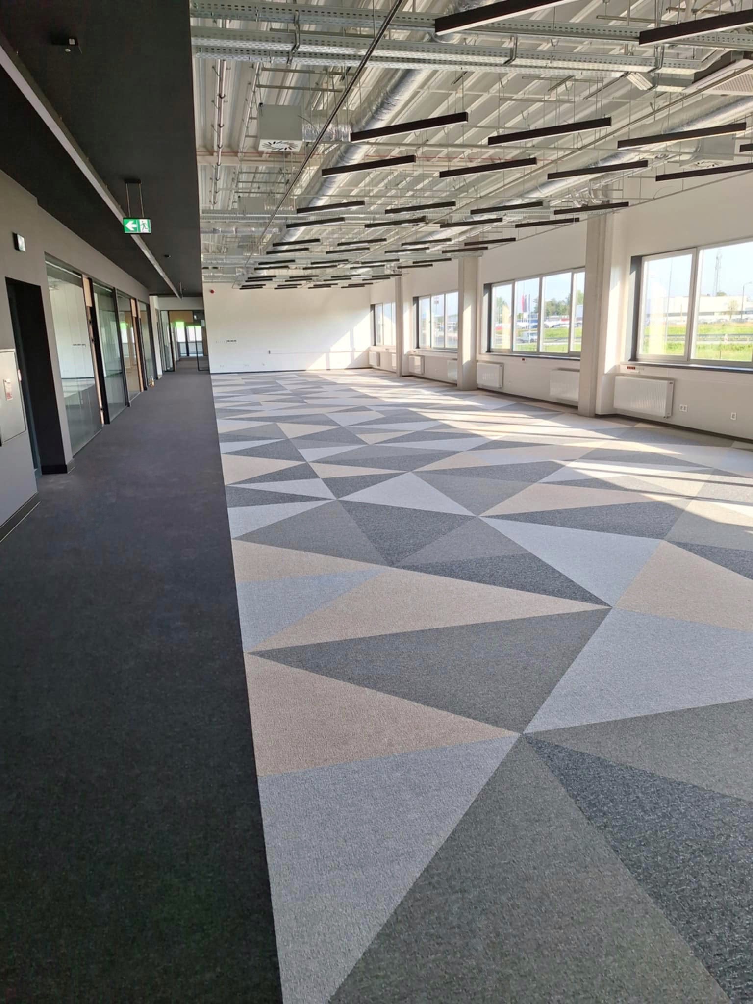 Przestronne biuro z wykładziną dywanową w geometryczne wzory, widok na okna i sufit z instalacjami. Część podłogi wyłożona ciemną wykładziną.