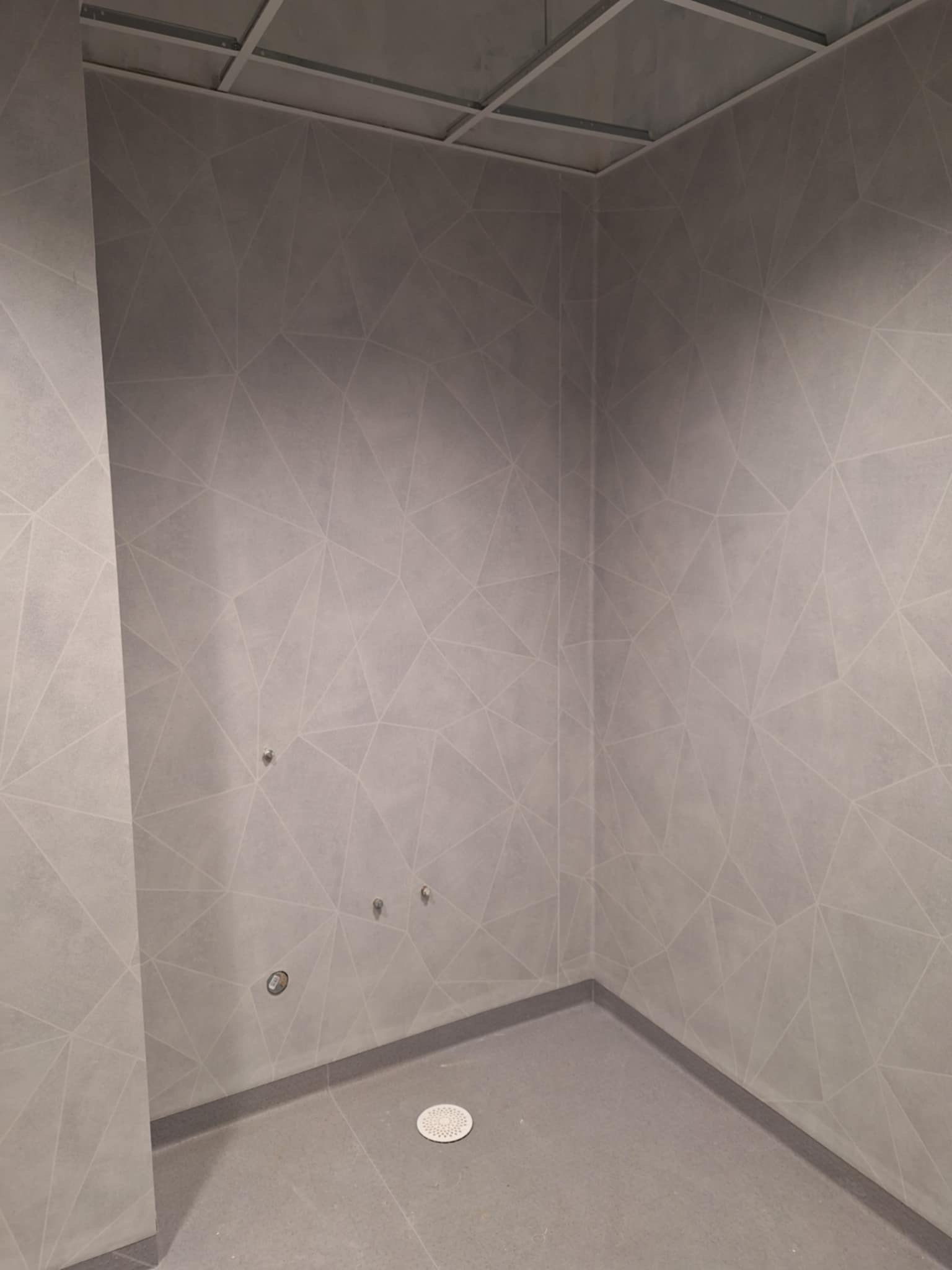 Wykończone wnętrze z szarą, geometryczną wykładziną ścienną i podłogową. Widoczne metalowe elementy sufitu podwieszanego i odpływ podłogowy.