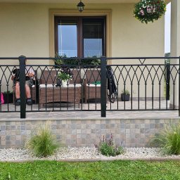 Zakład Usługowy Slusarstwo-Spawalnictwo PAVO - Czarna, metalowa balustrada z ozdobnymi elementami na tarasie domu jednorodzinnego, widok na ogród, osoba siedząca z psem na kolanach.