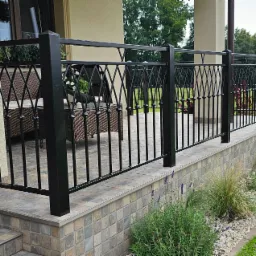 Czarna metalowa balustrada balkonowa z dekoracyjnym wypełnieniem, widok z zewnątrz, z fragmentem tarasu wyłożonego płytkami i roślinami wzdłuż balustrady.