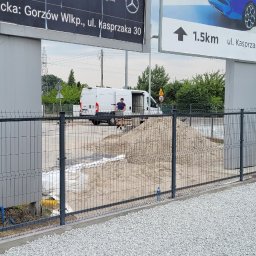 Zakład Usługowy Slusarstwo-Spawalnictwo PAVO - Szare ogrodzenie panelowe na betonowej podmurówce, z wysypanym białym żwirem i stertą piasku wewnątrz, biały bus z otwartymi drzwiami w tle, reklamy na słupach i toaleta przenośna po lewej stronie.