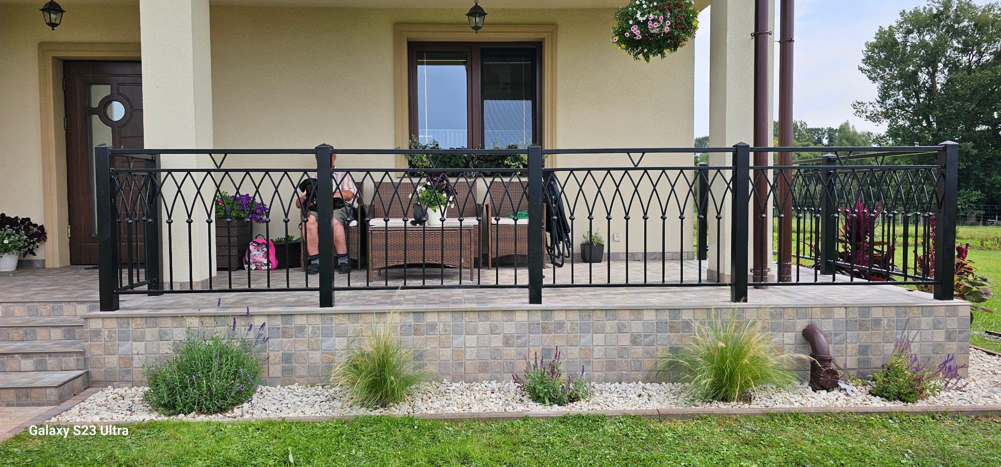 Czarna, metalowa balustrada z ozdobnymi elementami na tarasie domu jednorodzinnego, widok na ogród, osoba siedząca z psem na kolanach.