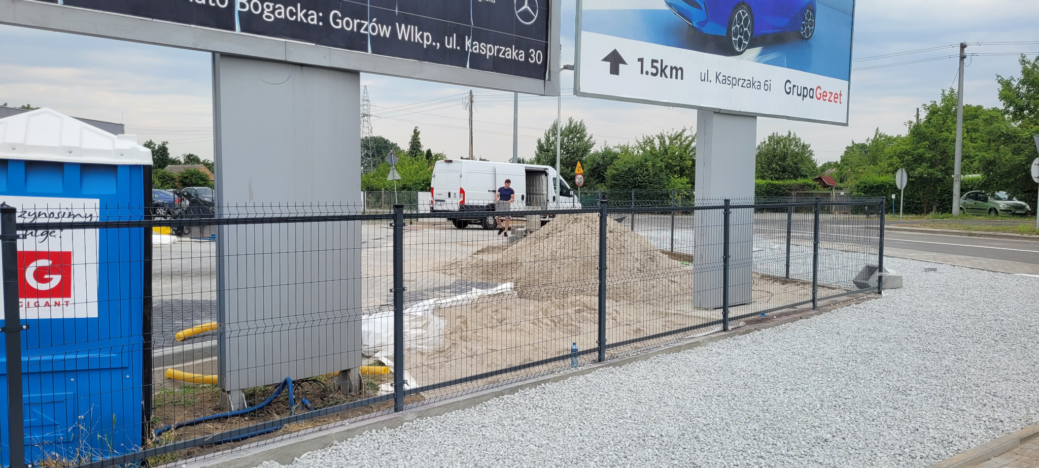 Szare ogrodzenie panelowe na betonowej podmurówce, z wysypanym białym żwirem i stertą piasku wewnątrz, biały bus z otwartymi drzwiami w tle, reklamy na słupach i toaleta przenośna po lewej stronie.