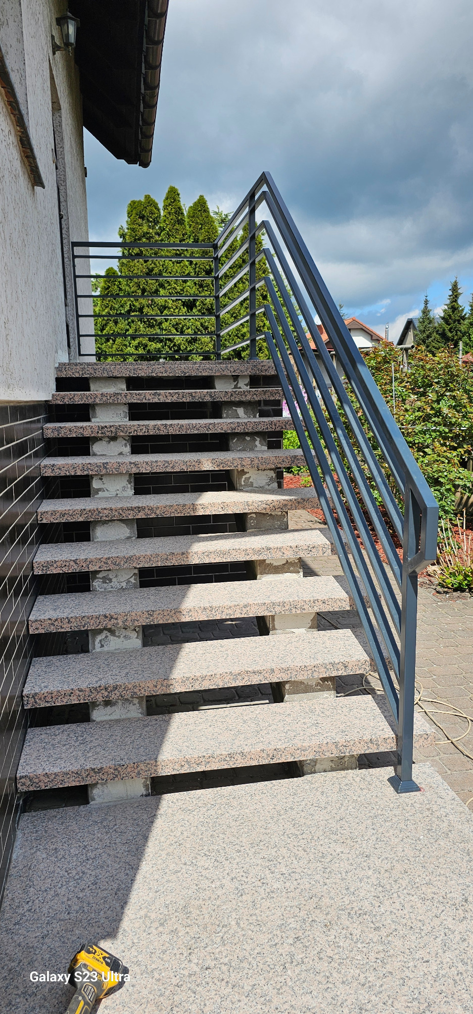 Zewnętrzne schody z granitu z nowoczesną, metalową balustradą w kolorze grafitowym, widok z dołu na tle zieleni.