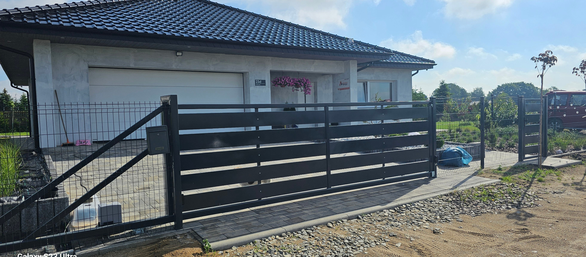 Nowoczesna posesja z czarną, poziomą bramą przesuwną i furtką z siatki panelowej, widok na dom z garażem i zagospodarowany ogród w tle.