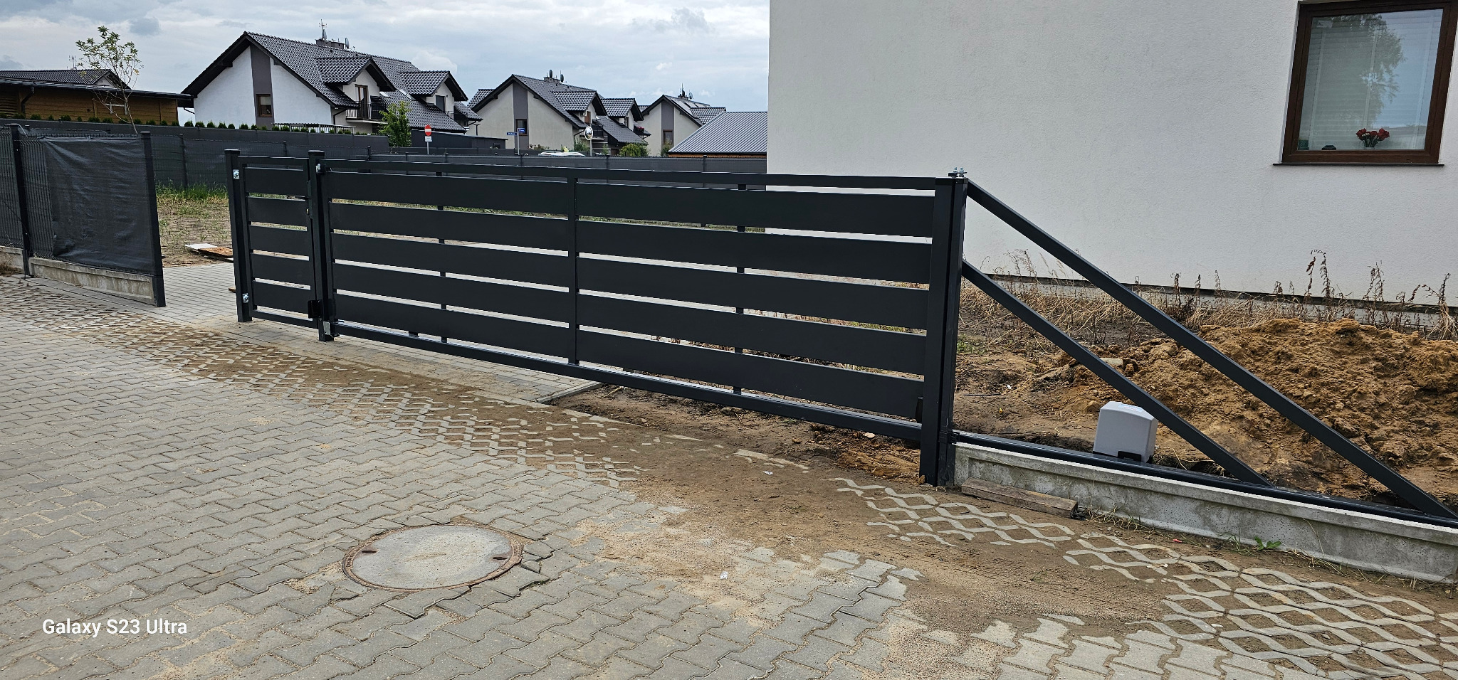 Nowoczesna, przesuwna brama wjazdowa z poziomych, metalowych profili, z widocznym napędem automatycznym, na tle kostki brukowej i domu jednorodzinnego.