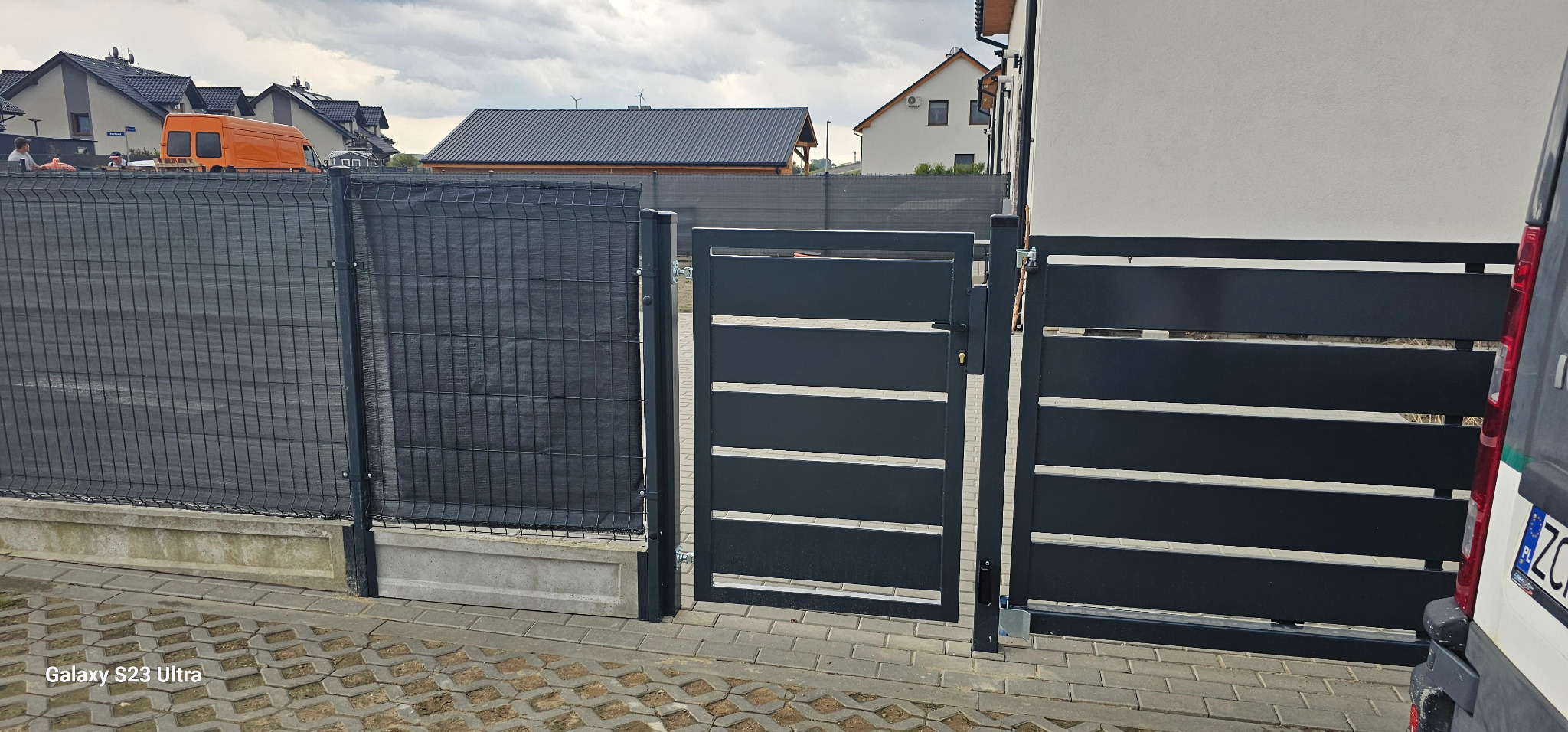 Nowoczesne, antracytowe ogrodzenie panelowe z przesłoną i brama wjazdowa z poziomymi profilami, widok z poziomu chodnika przed posesją.