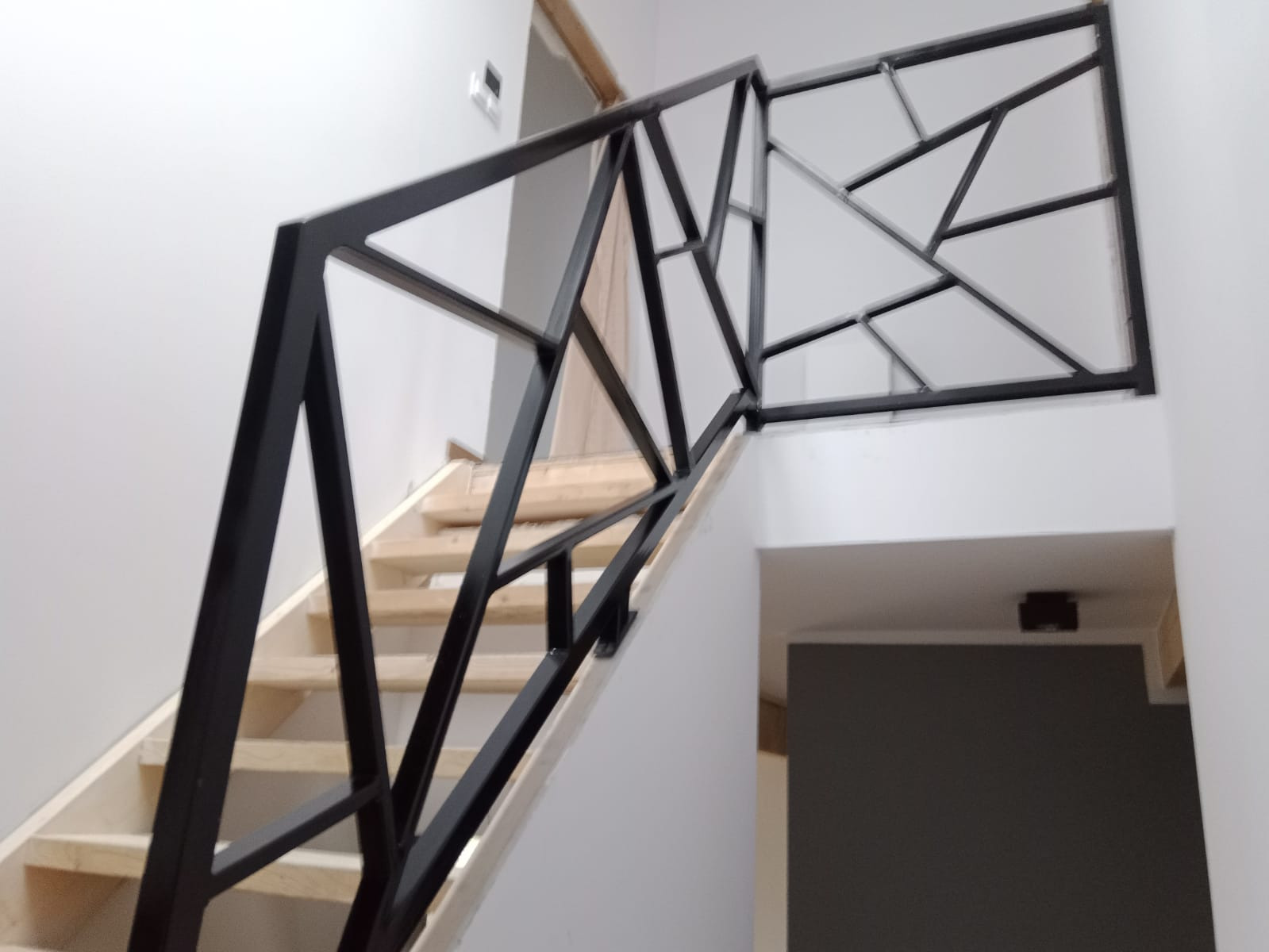 Czarna, metalowa balustrada schodowa o geometrycznym wzorze, zamontowana na drewnianych schodach w jasnym wnętrzu.