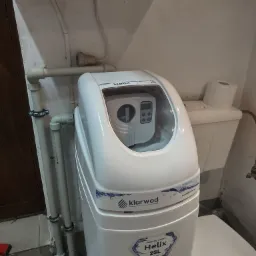 Biały zmiękczacz wody Klarwod Helix 25L w piwnicy, obok toaleta z rolką papieru, rury PCV z obejmami na ścianie, widoczne narzędzia na podłodze.