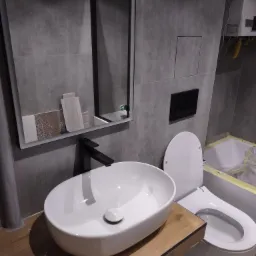 Nowoczesna łazienka w trakcie wykańczania: biała umywalka nablatowa z czarną armaturą na drewnianym blacie, wisząca toaleta, szare płytki wielkoformatowe na ścianach, podświetlane lustro z próbkami...