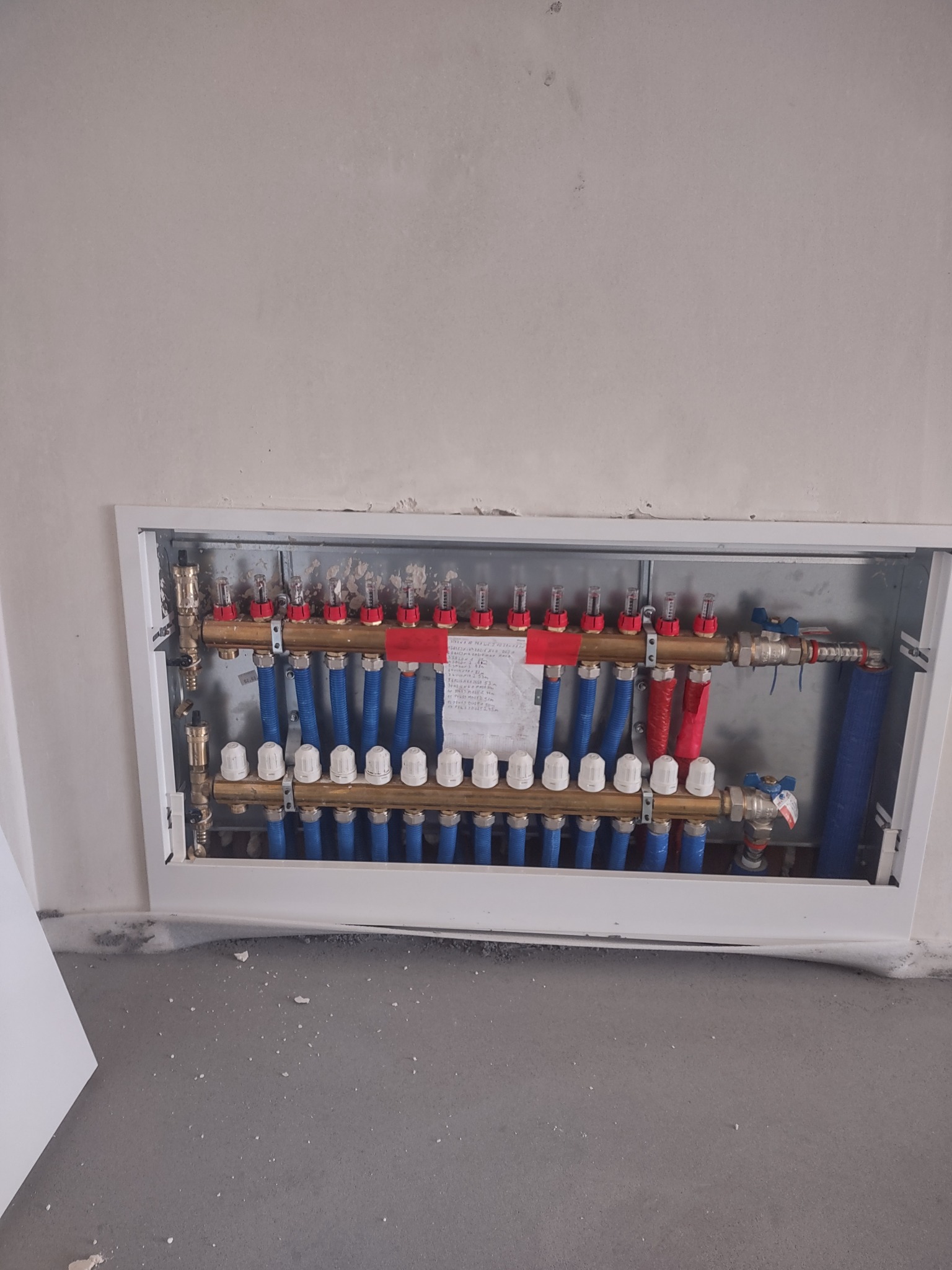 Rozdzielacz ogrzewania podłogowego w metalowej szafce, z widocznymi niebieskimi rurami, czerwonymi elementami i białymi pokrętłami, zamontowany w surowej ścianie.