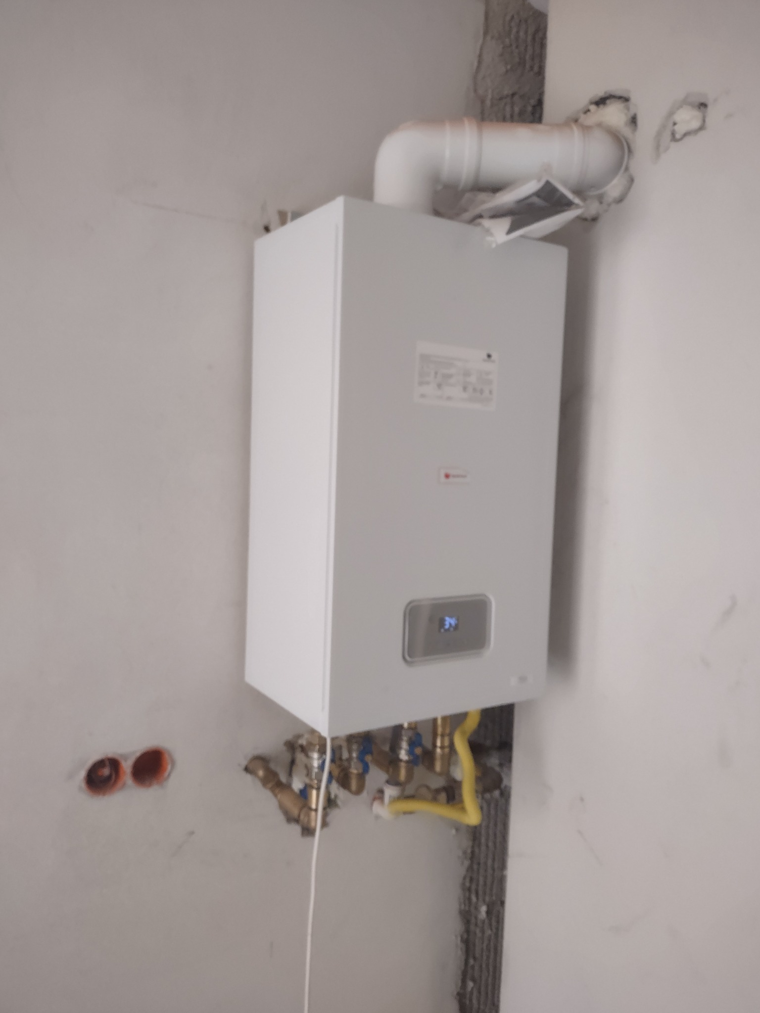 Biały, wiszący piec gazowy z podłączonymi rurami, zamontowany na surowej, otynkowanej ścianie w trakcie instalacji. Widoczne otwory w ścianie obok.