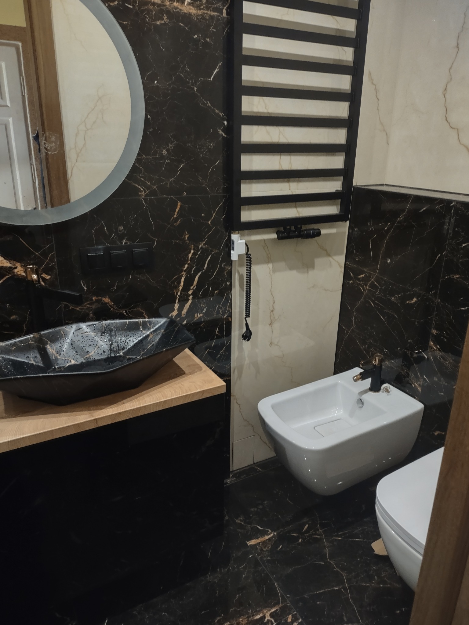 Nowoczesna łazienka z czarnymi marmurkowymi płytkami, umywalką nablatową w geometrycznym kształcie, białym bidetem z czarną armaturą i grzejnikiem drabinkowym w kolorze czarnym.