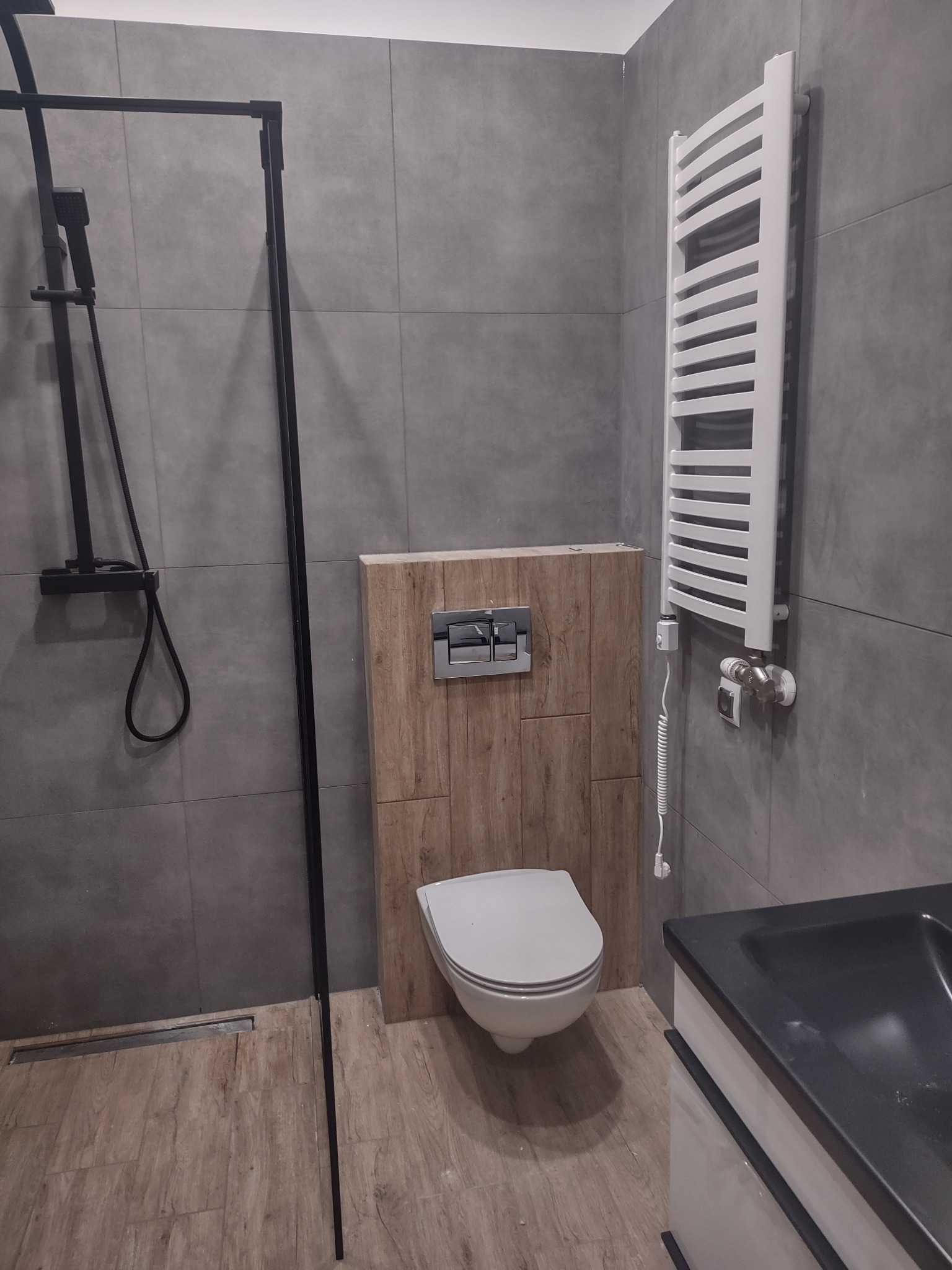 Nowoczesna łazienka z szarą glazurą, drewnopodobną zabudową stelaża WC, czarną kabiną prysznicową typu walk-in i białym grzejnikiem drabinkowym.