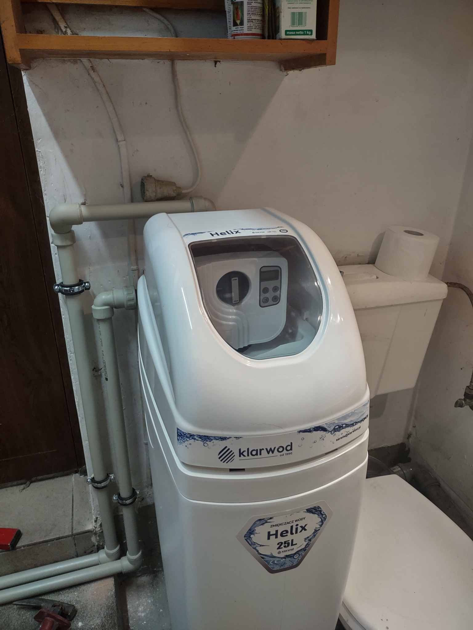 Biały zmiękczacz wody Klarwod Helix 25L w piwnicy, obok toaleta z rolką papieru, rury PCV z obejmami na ścianie, widoczne narzędzia na podłodze.