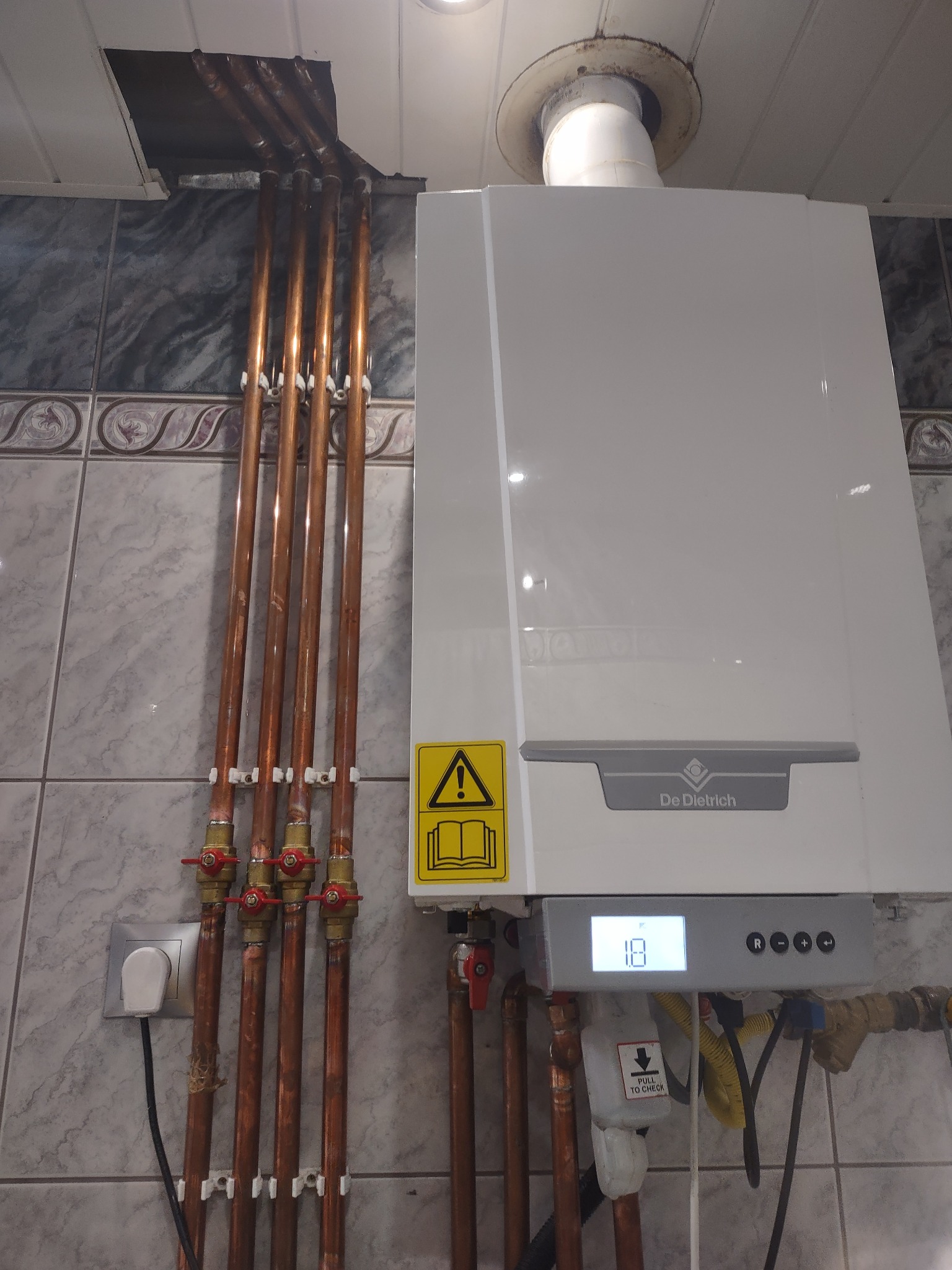 Biały piec gazowy De Dietrich zamontowany na ścianie z dekoracyjnymi płytkami, widoczne miedziane rury z zaworami, gniazdko elektryczne i wyświetlacz pieca wskazujący temperaturę 18 stopni.