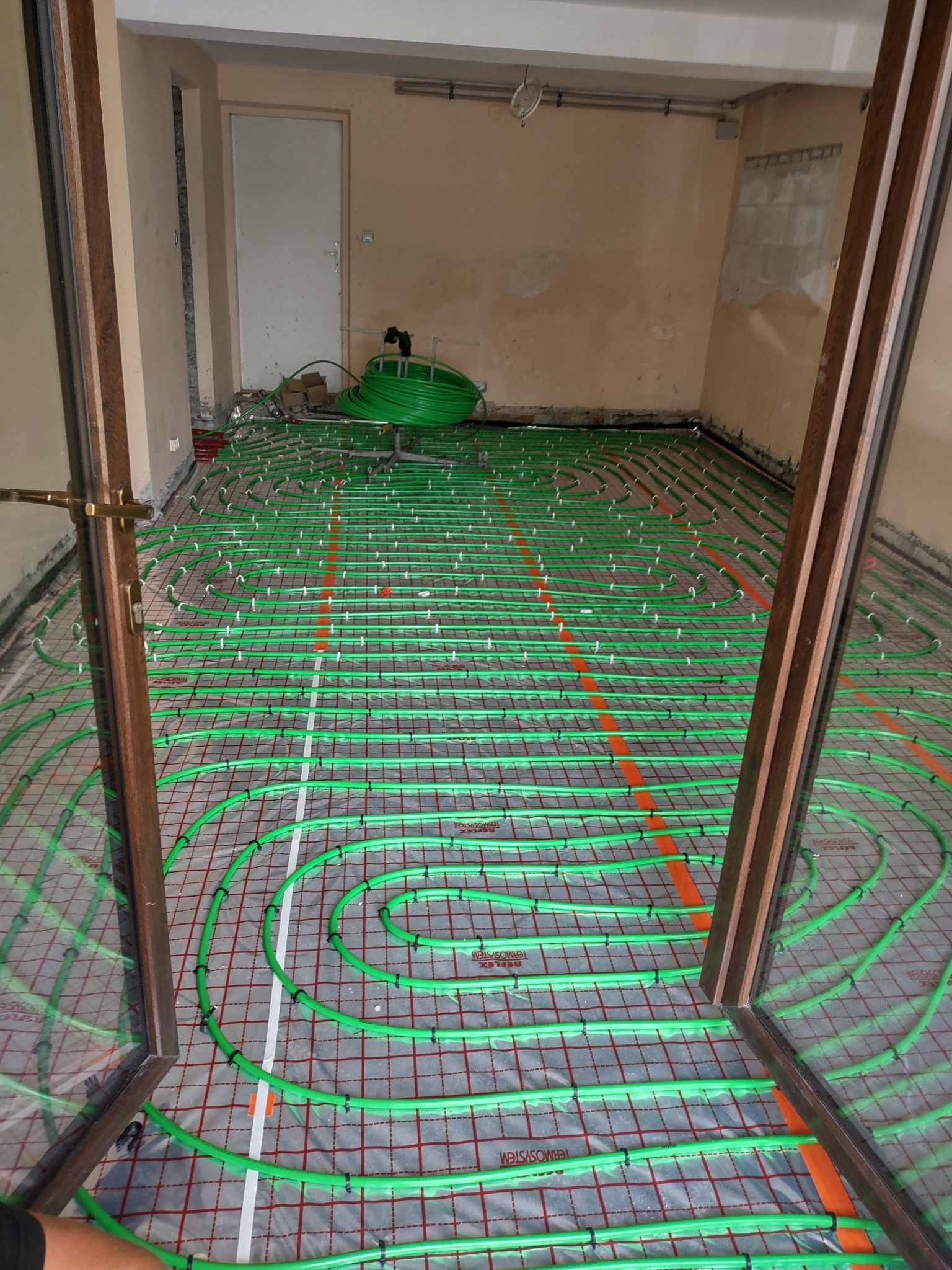 Zielone rury ogrzewania podłogowego rozłożone na folii z siatką, widok przez otwarte drzwi, w tle zwój rur.