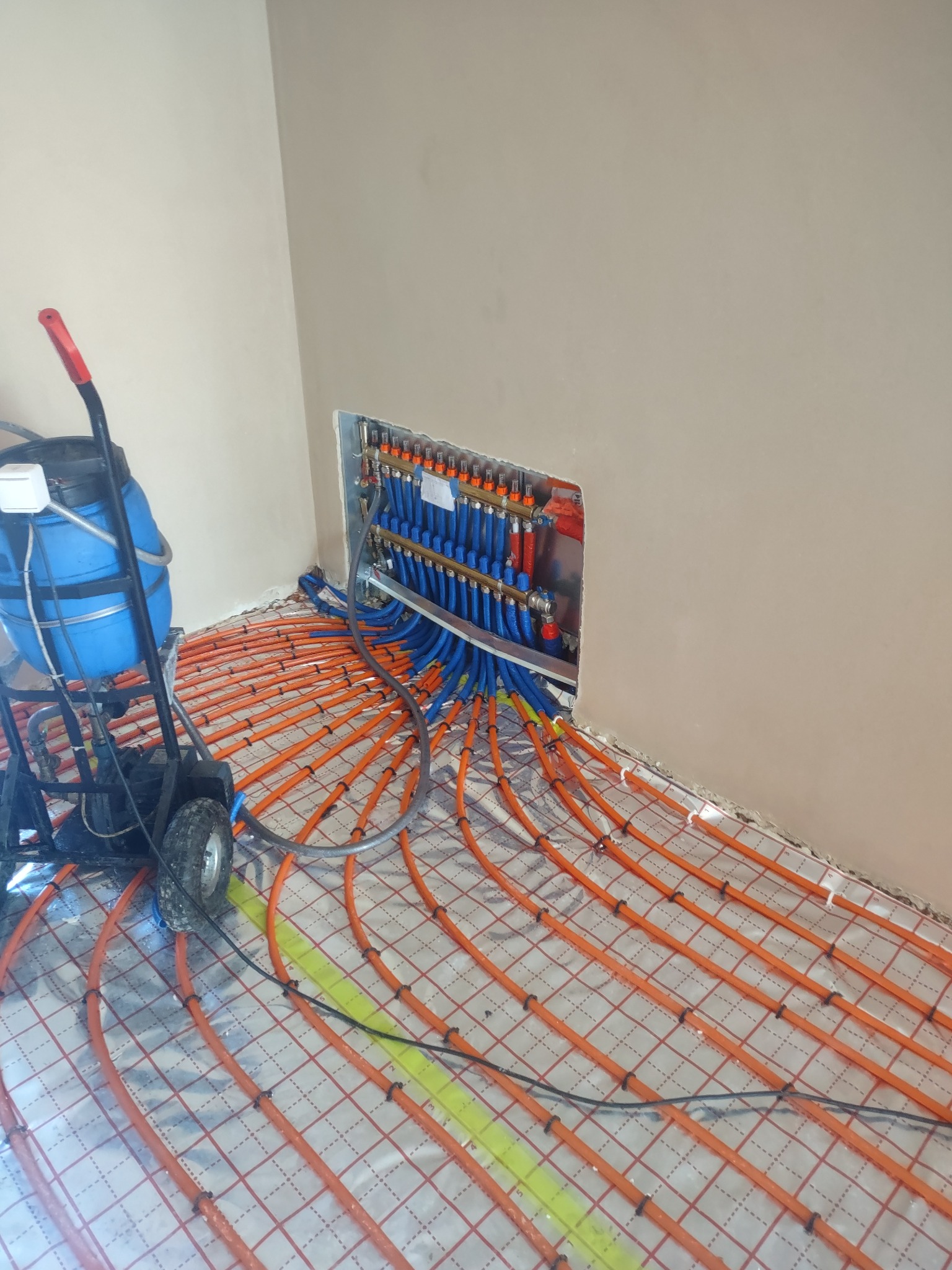 Rozłożona instalacja ogrzewania podłogowego z pomarańczowymi rurkami na folii aluminiowej, podłączona do rozdzielacza z niebieskimi rurami, widoczny wózek z niebieskim zbiornikiem.