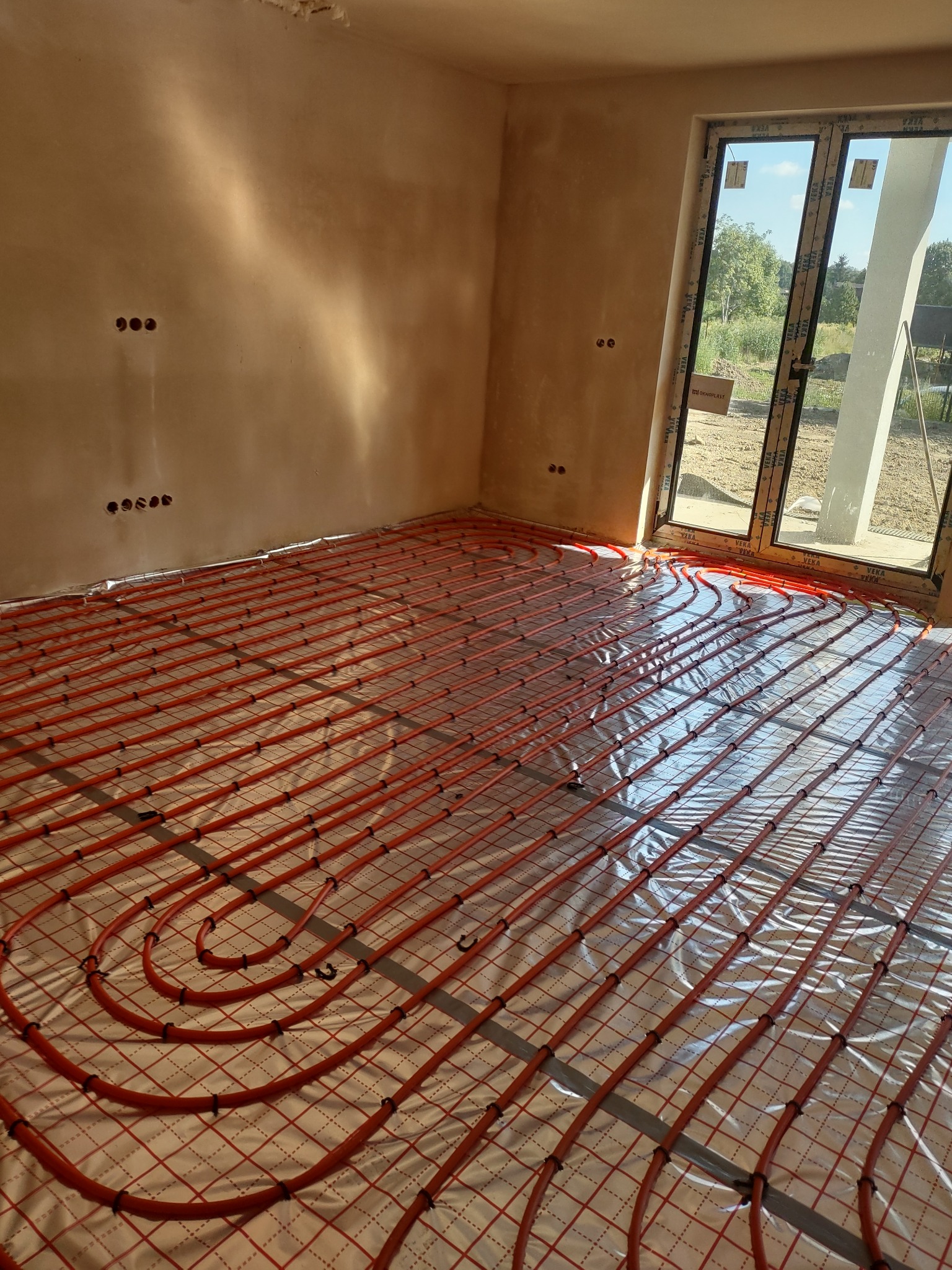Pomieszczenie w stanie surowym z widoczną instalacją ogrzewania podłogowego, rurki w kolorze pomarańczowym ułożone na folii z siatką, ściany w jasnym odcieniu beżu, okno z widokiem na zewnątrz.