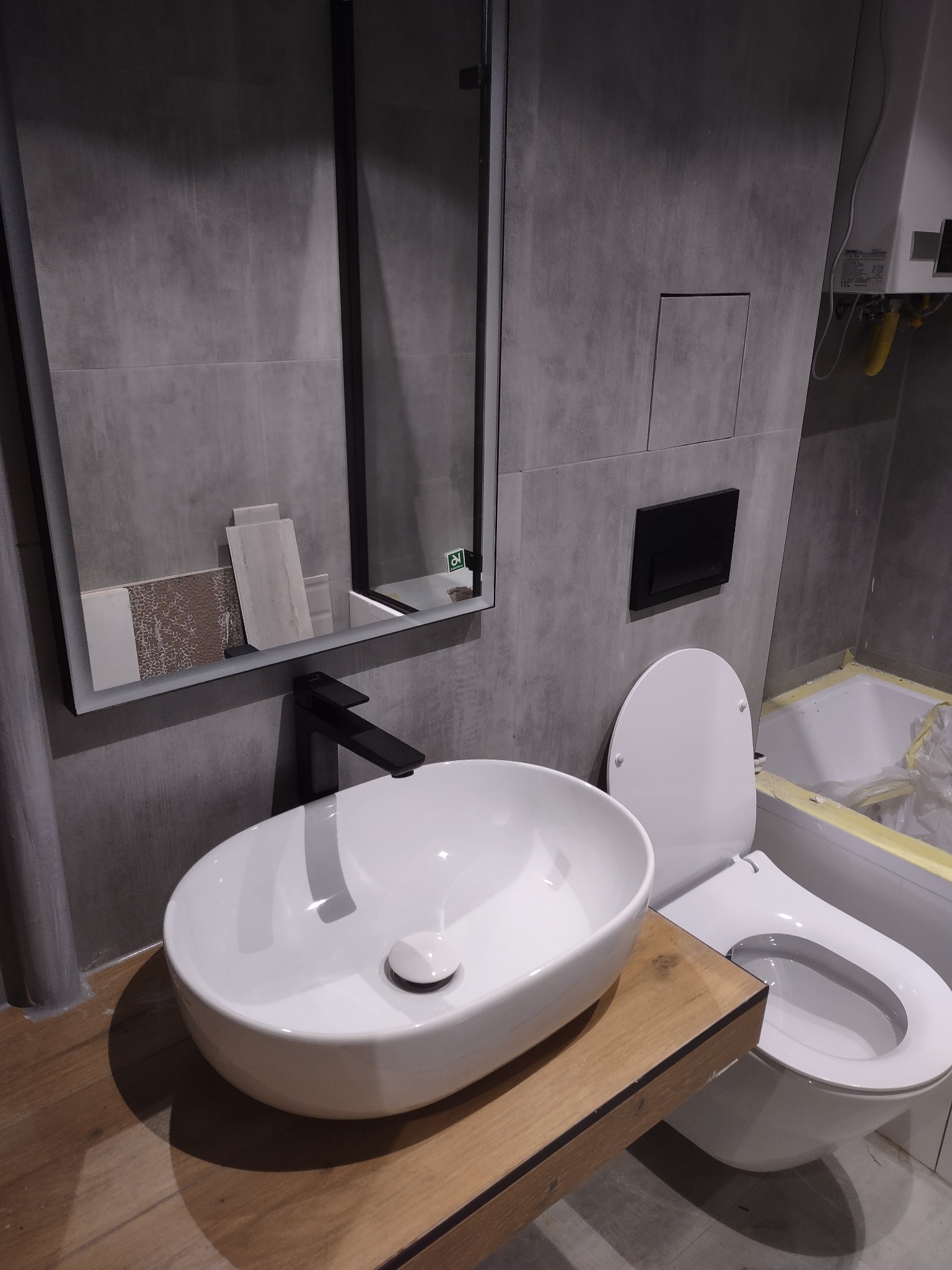 Nowoczesna łazienka w trakcie wykańczania: biała umywalka nablatowa z czarną armaturą na drewnianym blacie, wisząca toaleta, szare płytki wielkoformatowe na ścianach, podświetlane lustro z próbkami...