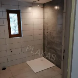 Wnętrze łazienki w trakcie remontu, widoczny brodzik, płytki w odcieniach szarości, okno z drewnianą ramą i przygotowane otwory na oświetlenie sufitowe.