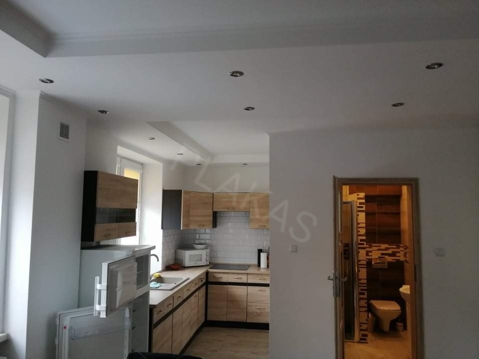 Wnętrze po remoncie: widok na kuchnię z drewnianymi szafkami i białą glazurą, sufit podwieszany z oświetleniem punktowym oraz otwarte drzwi do łazienki z brązową mozaiką.